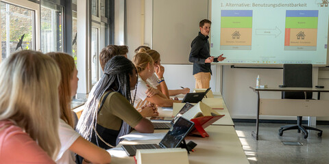 Es wird ein Dozent gezeigt, der im Lehrsaal vor der Tafel steht und etwas erklärt. De Studierenden blicken zu ihm nach vorne.