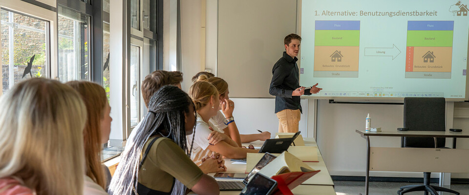 Es wird ein Dozent gezeigt, der im Lehrsaal vor der Tafel steht und etwas erklärt. De Studierenden blicken zu ihm nach vorne.