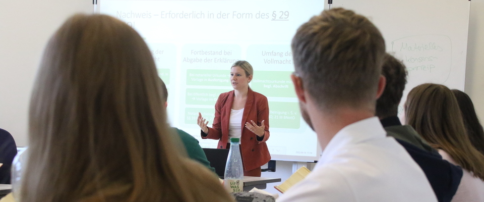 Es wird eine Dozentin vor der Studiengruppe in einer Lehrsituation gezeigt.